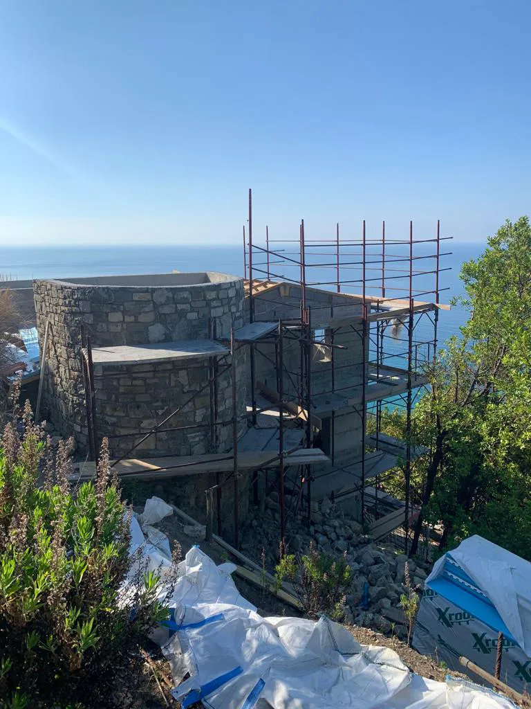 Edificio in costruzione a Mentone, con una sezione circolare rivestita in pietra e impalcature che ne avvolgono la struttura. Sullo sfondo, il mare blu e il paesaggio costiero della Costa Azzurra. L'immagine mostra l'architettura distintiva e la posizione privilegiata di un immobile con vista sul Mediterraneo.
