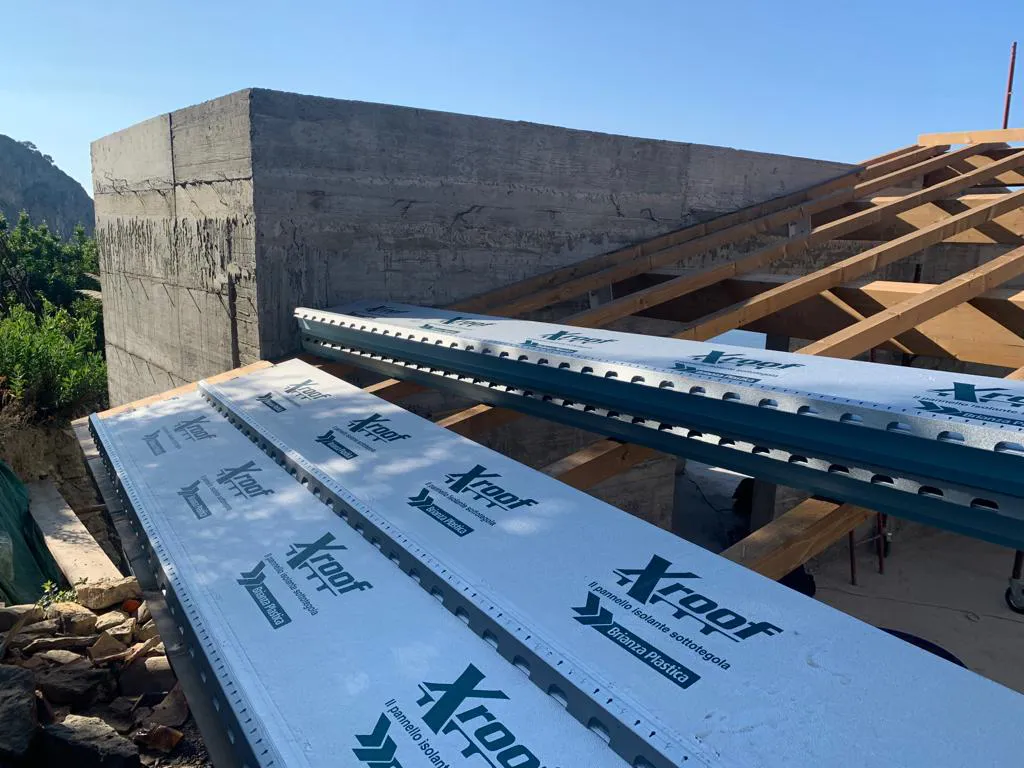 Pannelli isolanti "Xroof" con logo verde posizionati sulla struttura del tetto in legno di un edificio a Mentone. Sullo sfondo si vede una parete in cemento grezzo e alberi. L'immagine illustra la fase di installazione dei materiali isolanti ad alta efficienza energetica, essenziali per la costruzione moderna in Costa Azzurra.