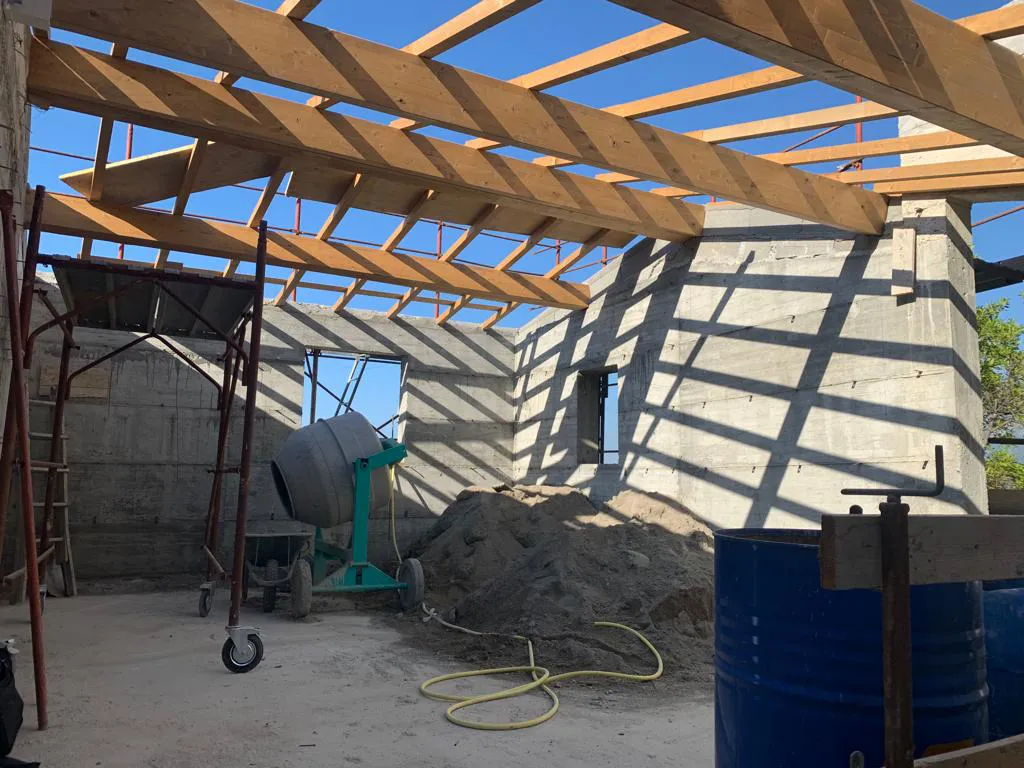 Interno di un edificio in fase avanzata di costruzione a Mentone, con il telaio del tetto in legno e pareti in cemento grezzo. Una betoniera e un cumulo di sabbia sono presenti, con ombre marcate. L'immagine illustra il progresso dei lavori all'interno del cantiere, inclusa la preparazione del solaio e del tetto per un immobile in Costa Azzurra.
