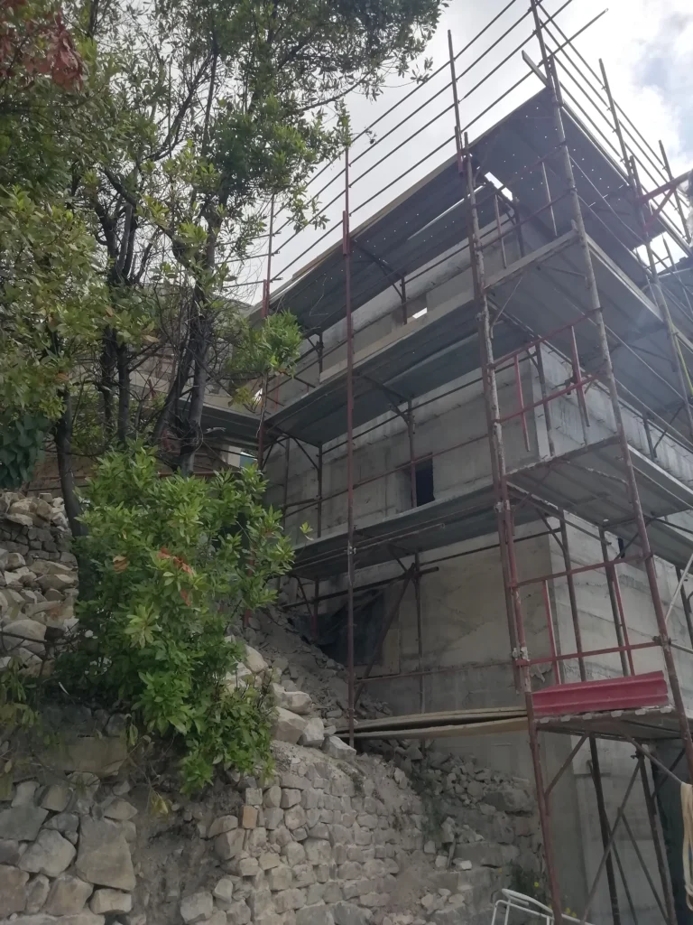 Edificio in fase di costruzione o ristrutturazione a Mentone, con impalcature complete che avvolgono la struttura in cemento grezzo. Alberi e vegetazione densa ai lati indicano un contesto naturale. L'immagine mostra l'avanzamento dei lavori edili nella Costa Azzurra.