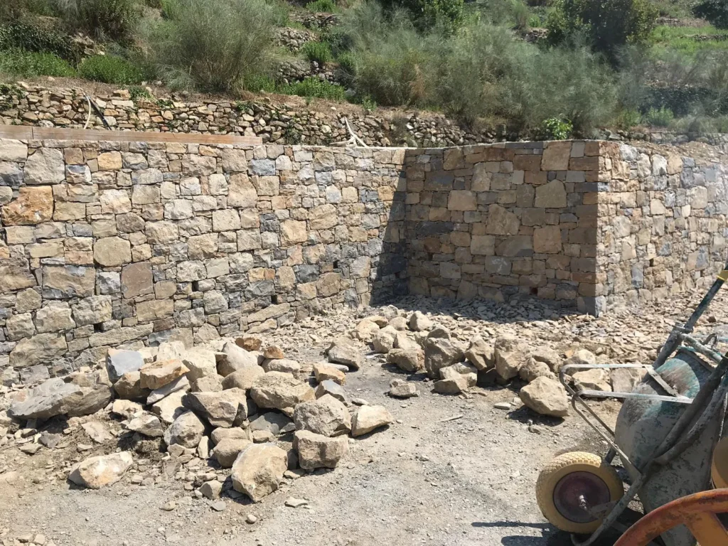 Cantiere per costruzione muro di contenimento in pietra naturale, con betoniera e pietre pronte per la posa. Realizziamo opere murarie solide e durevoli a Ventimiglia, Mentone e Costa Azzurra.